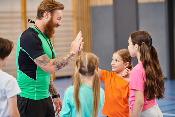 A male teacher stands in front of a group of diverse children, instructing them in a bright, lively classroom setting.