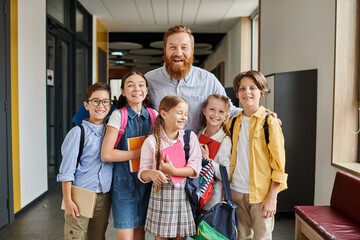 A dedicated teacher stands amongst a multicultural group of children in a vibrant school hallway,...