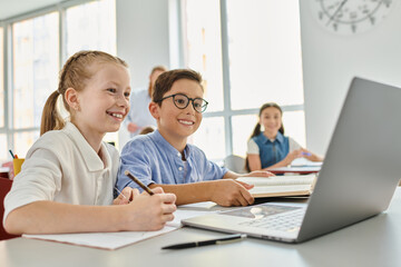 Obraz premium A group of children sitting at a table, focused on a laptop in a bright classroom.