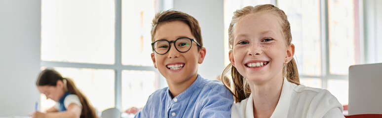 Two children, a boy and a girl, sitting side by side, engrossed in a conversation or activity in a vibrant classroom environment.
