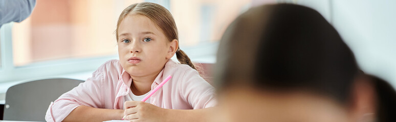 A young girl sits at a table, engrossed in her task, with a bored face expression in classroom...