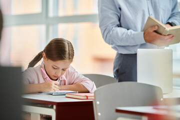 A young girl sits at her desk, engrossed in a book and using a calculator. The classroom is bright and lively, with a man teacher instructing her