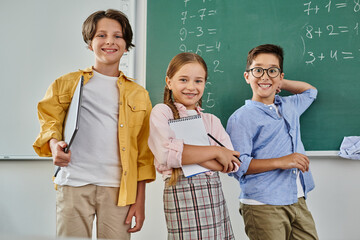 A group of children stand attentively in front of a chalkboard in a bright, lively classroom.