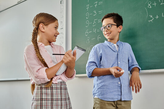 A boy and a girl stand in front of a blackboard in a bright classroom