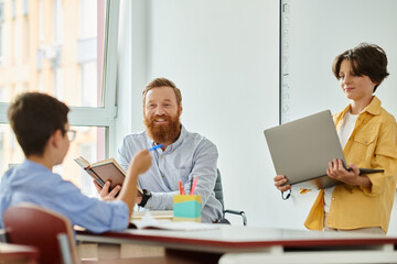 A group of individuals, including a teacher, engaged in a collaborative learning session around a table with laptops in a bright and lively classroom setting.