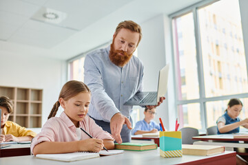 A male teacher assists a young girl with her homework in a vibrant classroom setting, surrounded by a diverse group of engaged students.