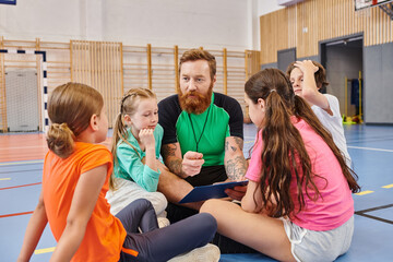 A male teacher sits on the floor surrounded by a diverse group of children in a bright and lively...