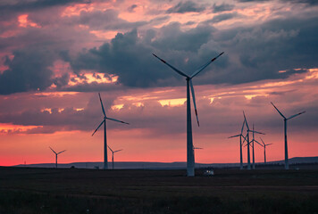 Obraz premium Multiple wind turbines silhouetted against a vibrant sunset sky. The turbines stand tall against the backdrop of orange and red clouds.