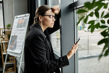 Middle-aged woman in glasses leans against window, looking at phone screen.