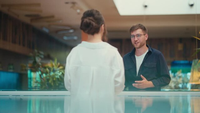 Cheerful Young Caucasian Man Coming at a Store Counter and Describing the Product He Needs to a Saleswoman. Concept of Retail, Sales, Shopping, Customer Service