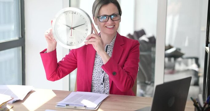 Positive female employee in glasses holds round wall clock in hands looking in camera and smiling. Time management at work in office slow motion
