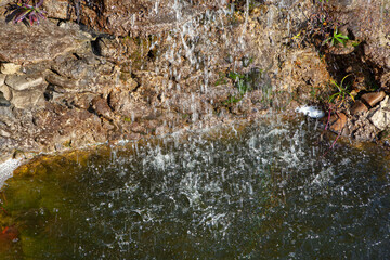 Small waterfall is flowing into a pond. The water is murky and the rocks surrounding the pond are grey