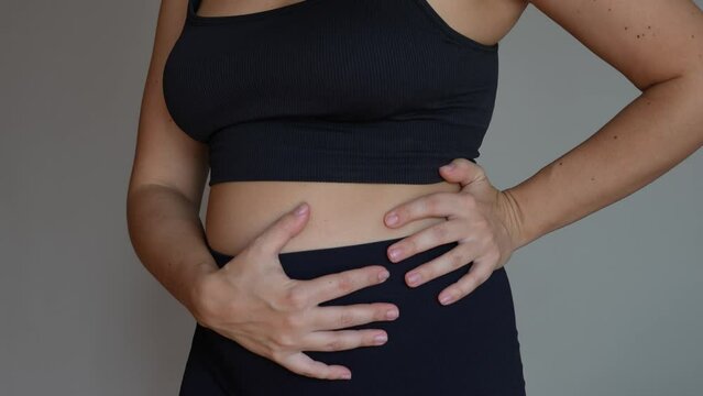 A young woman holding her belly with her hands on grey background. Stomach ache, bloating, flatulence, abdominal distension, colic, heaviness, digestive disorders. Overeating, overweight, food issues