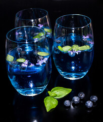 Three glasses of vibrant blue blueberry flavoured Gin and Tonnic, garnished with basil leaves, rosemary flowers and extra blueberries