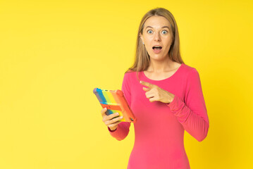 Shocked excited young blond woman with tablet pc isolated on yellow background. Surprised woman with open mouth in red fitted dress pointing to tablet pc holding in hand