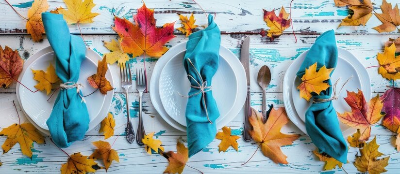 A fall themed table setting featuring vibrant leaves a teal napkin and silverware on a white shiplap board ideal for a copy space image