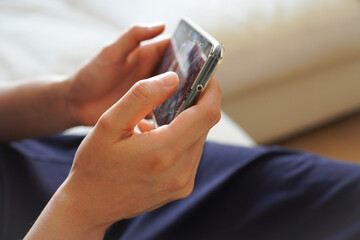 ソファーに座って、スマホ(スマートフォン)を横持ちして動画を見る若い女性