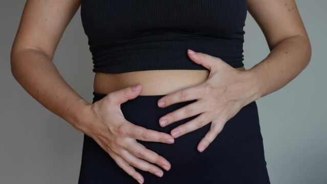 A young woman holding her belly with her hands on a grey background. Stomach ache, bloating, flatulence, abdominal distension, colic, heaviness, digestive disorders. Overeating, overweight