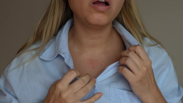 Panic attack. Young blonde woman keeping her hand on her chest in pain taking deep breaths of air unbuttoning the top button of her shirt on grey background. Chest pain, lack of breath
