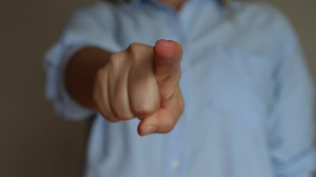 Cropped shot of a young woman in a blue shirt pointing forward into the screen with her hand on a beige background. Pointing finger gesture. The sign that means I choose you. Changing focus