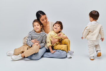 A young Asian mother sits on the ground with her children in a studio setting against a grey background, bonding closely.