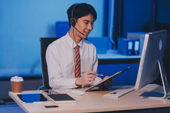 Happy smiling Asian man work at call center service desk consultant with teammates at night, customer service executive with microphone headset use computer for supporting in late night office.