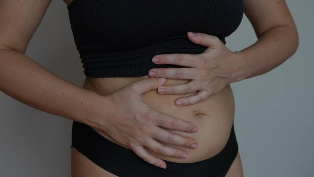 Stomach ache, bloating, flatulence, abdominal distension, colic, heaviness, digestive disorders. A young woman holding her swollen belly with her hands on dark grey background. Overeating, overweight