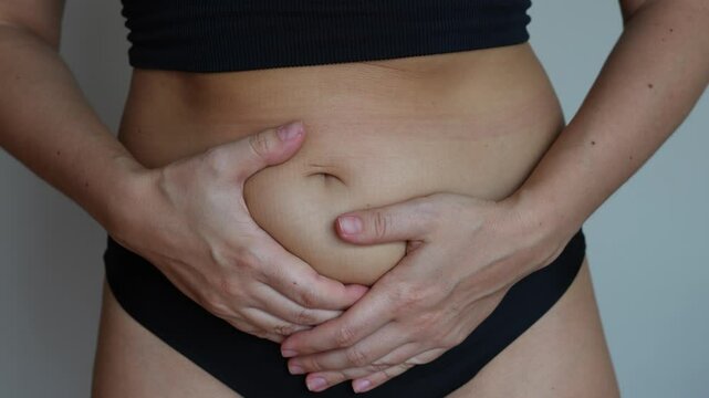 Cropped shot of a young woman holding her swollen lower abdomen with hands suffering from painful periods on dark grey background. Gynecological problems, genital tract infections, inflammation