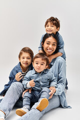 A young Asian mother and her little sons playfully stack on top of each other, all wearing denim clothes in a grey studio.