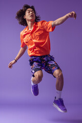 A full body shot of an asian male model wearing white socks, a red shirt and black shorts with a patterned design jumping in the air wearing purple shoes, against a purple background with studio light