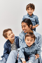 A group of young children, a mother, and her sons, all dressed in denim clothes, sitting next to each other in a grey studio.