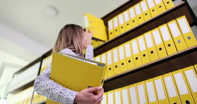 Secretary reaches over to upper shelf to take ring binder for paperwork. Organized yellow folders with archive materials in office slow motion