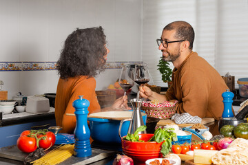 A diverse Hispanic couple smiles and toasts with glasses of wine, in the kitchen at home, waiting for dinner to finish cooking