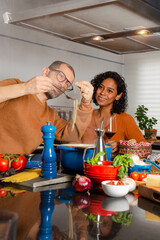 A Hispanic couple is in kitchen preparing an anniversary dinner. The man checks the pasta, while the woman, with a glass of wine, smiles