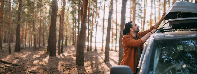 A young man opens the upper tourist trunk of a car in a pine forest on the shore of a lake at sunset. © Anna