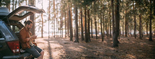 A young man with a phone in his hands next to a car in a pine forest on the shore of a lake at sunset. A tourist with a smartphone and an SUV  on a forest road © Anna