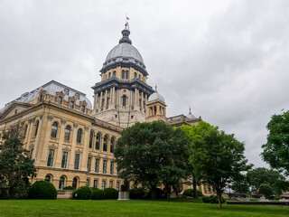 Front side view of the Illinois State Capitol Building in Springfield, IL, USA. Cloudy overcast skies overhead. Springtime scene with a lush green lawn and full trees.