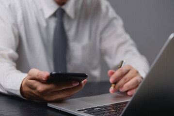 Businessman holding smartphone, using laptop. Managing tasks, modern office setup. Focus on digital communication, business operations. Professional workspace, efficient planning.