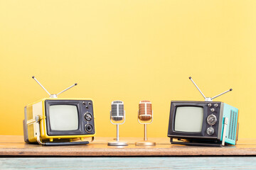 Old TVs, golden and silver microphones on wooden table front yellow background. Retro style filtered photo