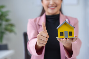 Real estate agent holding a small yellow house model and giving a thumbs up, indicating a promising investment opportunity