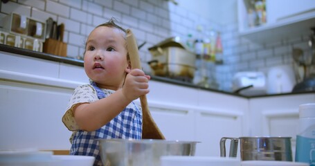 Adorable toddler child in a modern kitchen holding a wooden spoon mixing flour in bowl for bake a cake, engaging in playful cooking activities.