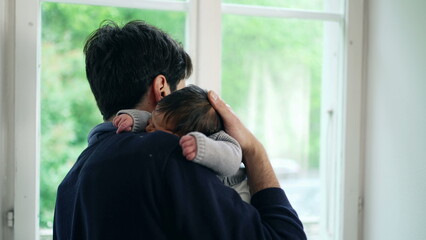 Father cradling newborn baby in arms, looking out window, tender moment, daytime, family love, natural light, indoor scene, parent-child bond, serene and peaceful, young fatherhood