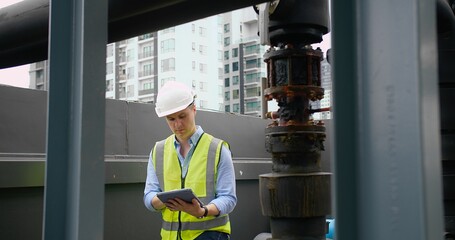 A Engineer man looking inspecting maintenance insulated pipelines valve pump control on the roof at an industrial site. He is wearing a hard hat and safety vest