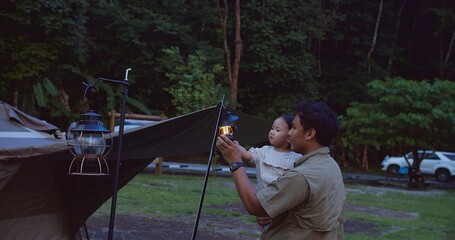 A father holding his child Enjoying Camping Trip in Forest with illuminated by lanterns at dusk, The scene captures a serene and bonding moment in nature.