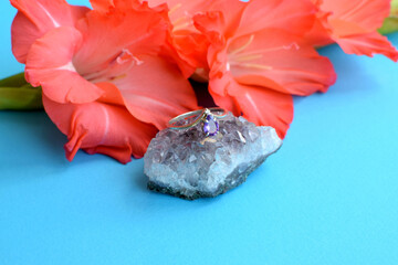 A silver ring with natural amethyst lies on an amethyst druse and a pink gladiolus flower.