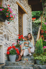 Mom and daughter in the garden with flowers. A European house with a courtyard, a European village. Mom, a young girl with long blonde hair, in a white dress and a scarf