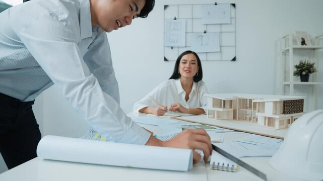 Skilled caucasian interior designer working with house model while discussing with asian architect engineer about house plan on meeting table with blueprint and house model scatter around. Immaculate.
