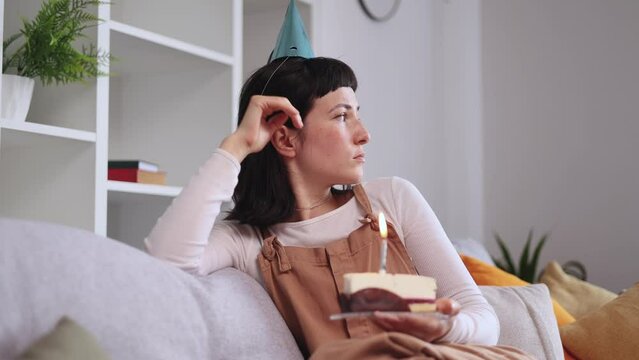 Upset woman in birthday party hat celebrating alone looking at cake with candle at home Pretty female feeling sad because everybody forgot about her event indoors Bad birthday mood concept