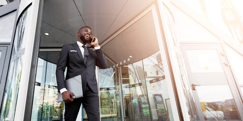 Happy Afro Business Guy Having Phone Conversation Against Entrance To Office Building. Copy Space