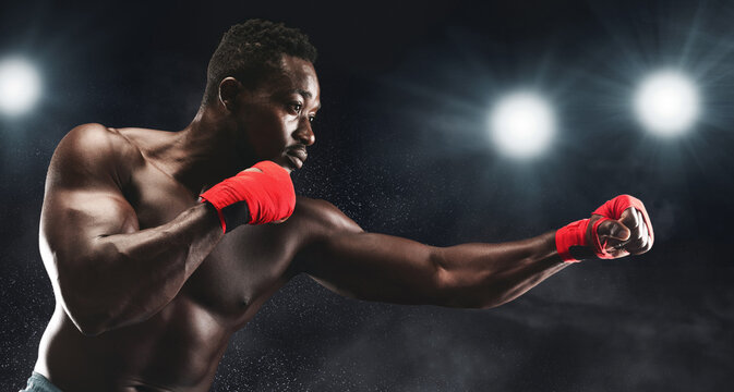 A muscular boxer with red wraps on his hands practices his punch in a dark boxing ring under bright lights. - Powered by Adobe